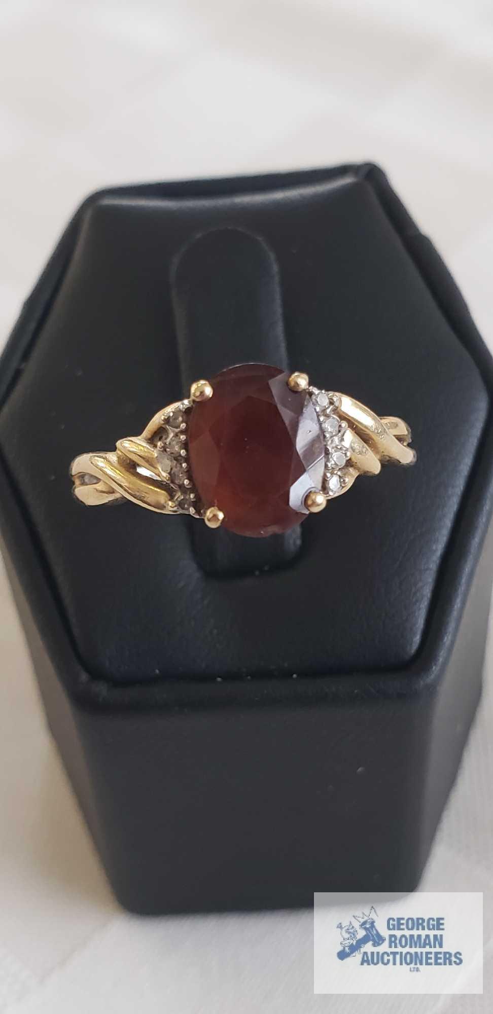 Gold colored ring with a large dark red gemstone, clear gemstones on side, twisted band, marked 14K,